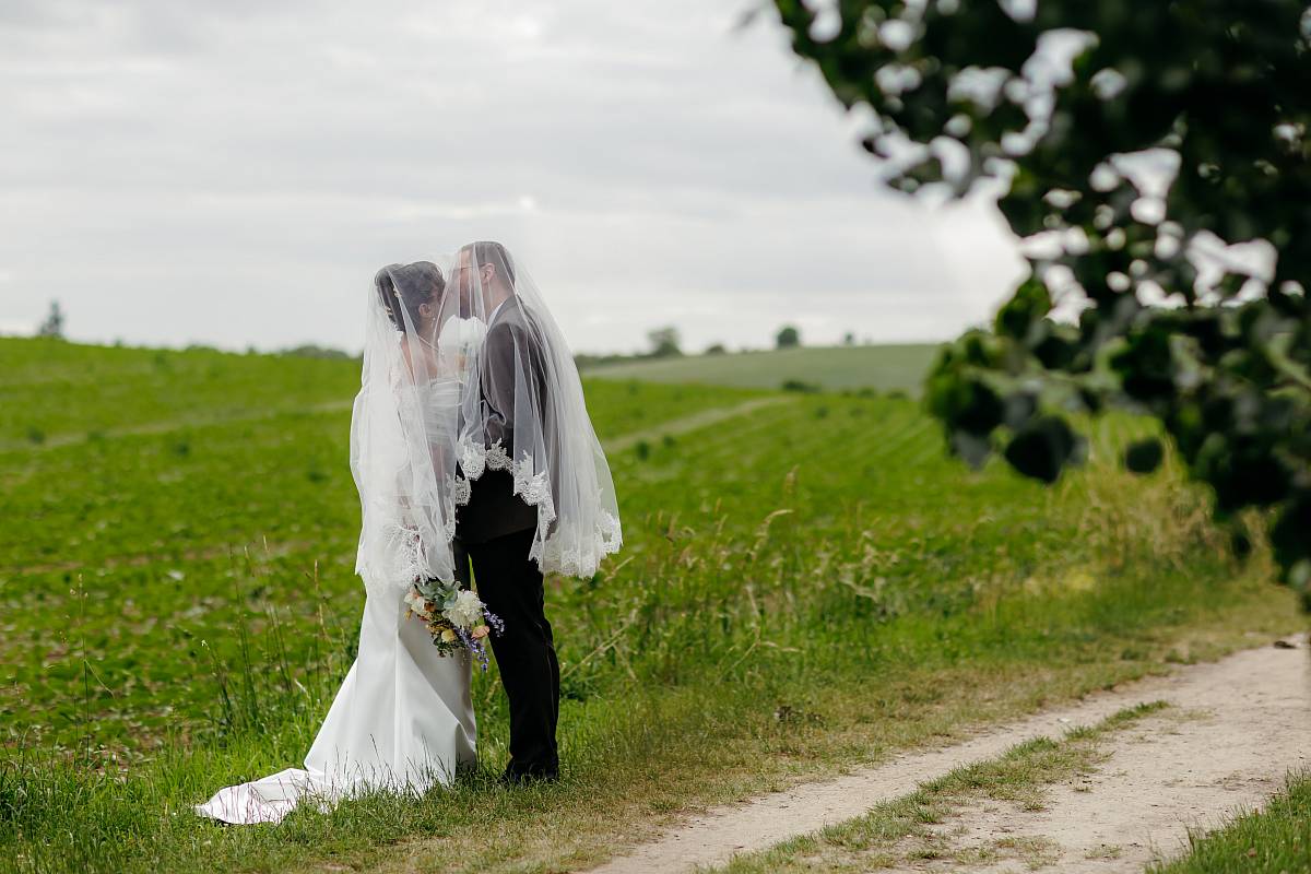 Les mariés s'embrassent sous le voile dans les champs derrière la ferme du prieuré de Rennemoulin.